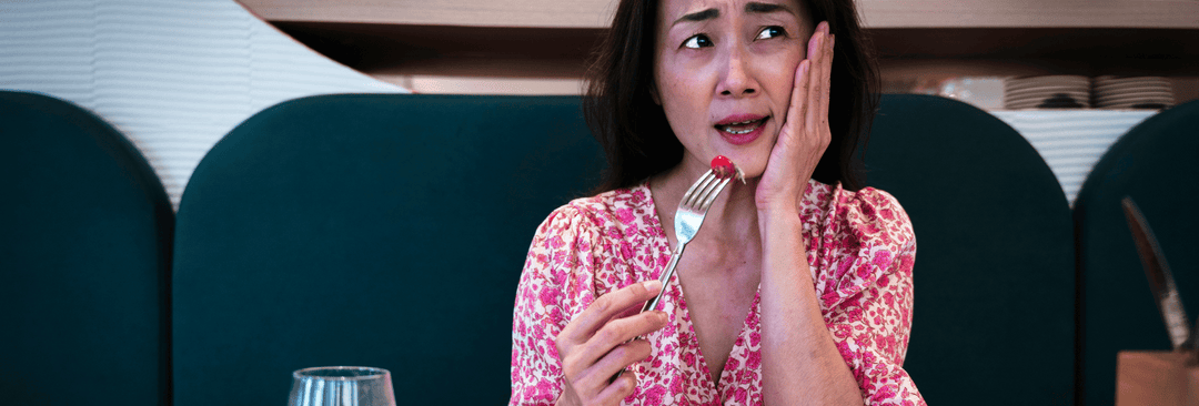 Concerned woman sitting at a restaurant table, holding a fork with a cherry tomato, looking distressed and touching her cheek, reflecting discomfort or a negative reaction to food.